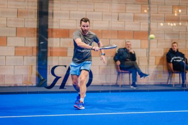 Riga, Letonya - 5 Nisan 2025 - Salıncakta sallanan erkek padel oyuncusu, mavi bir kortta gri gömlek ve mavi şort giyerek, kapalı bir padel turnuvası sırasında yoğun bir şekilde odaklanır..