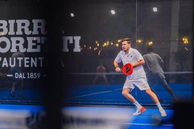 Riga, Letonya - 5 Nisan 2025 - Beyazlı erkek padel oyuncusu yoğun bir maç sırasında bir padel turnuvasında mavi bir kortta parlak kırmızı raket tutarak şut atmaya hazırlanıyor..