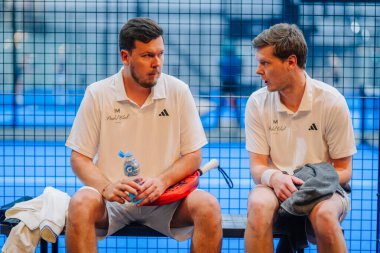 Riga, Letonya - 5 Nisan 2025 - Beyaz üniformalı iki erkek Padel turnuvasının ortasında elinde raket ve su şişesiyle bankta mola verdi..