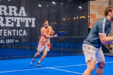 Riga, Letonya - 5 Nisan 2025 - İki erkek Padel oyuncusu çiftler maçında yarışıyor; biri kapalı alanda yapılan bir padel turnuvası sırasında topa yoğun bir şekilde vuruyor.