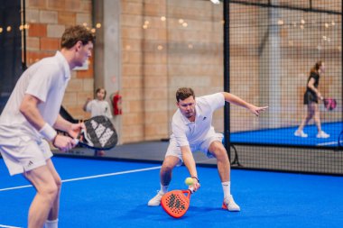 Riga, Letonya - 5 Nisan 2025 - Beyaz elbiseli iki erkek padel oyuncusu bir noktada hareket halindeler, biri kapalı alanda yapılan hızlı bir Padel turnuvasında topa doğru geriliyor.