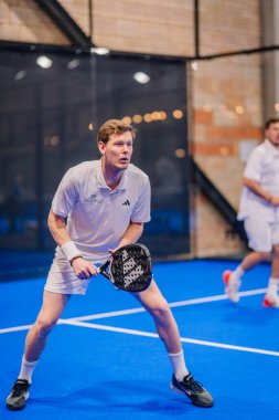Riga, Letonya - 5 Nisan 2025 - Beyaz spor giyim üzerine odaklanmış padel oyuncusu, kapalı alandaki bir sahada düzenlenen yoğun bir padel turnuvası sırasında atış yapmaya hazırlanıyor..