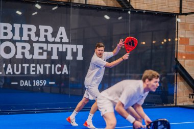 Riga, Letonya - 5 Nisan 2025 - Beyaz kıyafetli iki erkek padel oyuncusu bir puan için hazırlanıyor, biri kırmızı raketle, kapalı alanda oynanan bir Padel turnuvası sırasında.