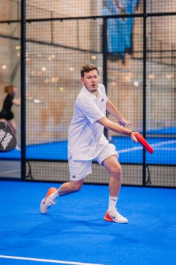 Riga, Letonya - 5 Nisan 2025 - Tamamen beyaz giysili bir erkek padel oyuncusu, kapalı alandaki bir sahada düzenlenen bir turnuva sırasında şutunu geri atmaya odaklanıyor..