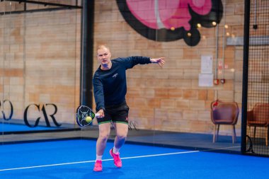 Riga, Letonya - 5 Nisan 2025 - koyu renk kazaklı ve pembe ayakkabılı odaklanmış bir erkek Padel turnuvası sırasında elini uzatır..