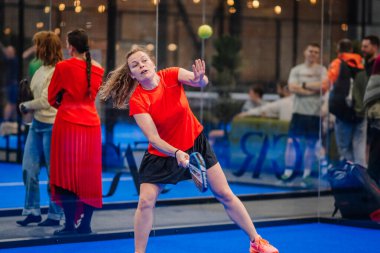 Riga, Letonya - 5 Nisan 2025 - Kırmızı tişörtlü bayan padel oyuncusu Padel turnuvası sırasında güçlü bir forehand atışı yapıyor..