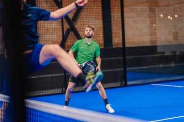 Riga, Letonya - 5 Nisan 2025 - Yeşil tişörtlü erkek padel oyuncusu Padel turnuvası sırasında oyuna odaklanırken takım arkadaşı topu ağdan geçirmek için zıplıyor.