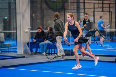 Riga, Letonya - 5 Nisan 2025 - Padel turnuvası maçında arka planda seyirciler ve diğer oyuncuların yer aldığı düşük vuruş yapan kadın oyuncu.