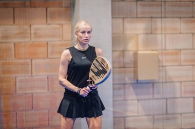 Riga, Letonya - 5 Nisan 2025 - Siyah spor giyim giymiş kadın padel oyuncusu elinde raket, odaklanmış ve kapalı sahadaki bir padel turnuvasının bir sonraki aşaması için hazır.