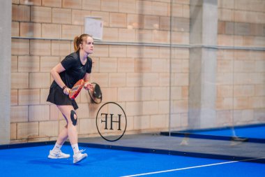 Riga, Letonya - 5 Nisan 2025 - Siyah spor giyim üzerine odaklanmış bayan padel oyuncusu kapalı mavi bir kortta düzenlenen bir padel turnuvası sırasında servis almaya hazırlanıyor.
