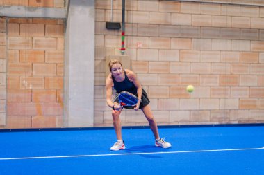 Riga, Letonya - 5 Nisan 2025 - Odaklanmış kadın padel oyuncusu, bir padel turnuvası maçında kapalı mavi bir sahada ters vuruş yapmaya hazırlanıyor..