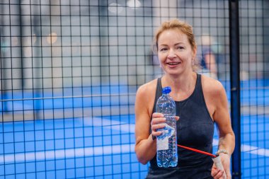 Riga, Letonya - 5 Nisan 2025 - Gülümseyen bayan padel oyuncusu, kapalı mavi bir kortta düzenlenen bir turnuva sırasında bir su şişesi ve padel raketi tutuyor.