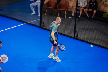 Riga, Letonya - 5 Nisan 2025 - Erkek oyuncu, arka planda izleyicilerin izlediği kapalı bir sahadaki bir Padel turnuva maçında forehand atışına hazırlanıyor..