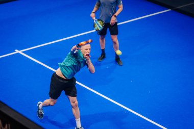 Riga, Letonya - 5 Nisan 2025 - Padel turnuvası sırasında iki erkek oyuncu, canlı bir mavi sarayda güçlü bir dönüş için ileriye doğru sıçrıyorlar..