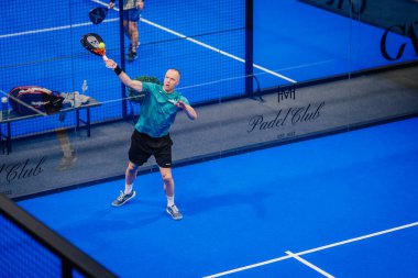 Riga, Letonya - 5 Nisan 2025 - Padel turnuvasındaki bir iç saha maçında parlak mavi döşemeli bir forehand vuruşu yapan erkek atlet.