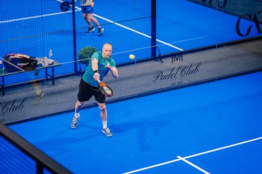 Riga, Letonya - 5 Nisan 2025 - Padel turnuvasındaki bir iç saha maçında parlak mavi döşemeli bir forehand vuruşu yapan erkek atlet.