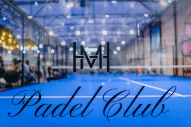 Riga, Letonya - 5 Nisan 2025 - Padel Club logosunun kapalı bir sahada çekilmiş yakın çekimi ve modern bir spor salonunda düzenlenen bir padel turnuvası sırasında arka planda oyuncular bulanıklaştı.