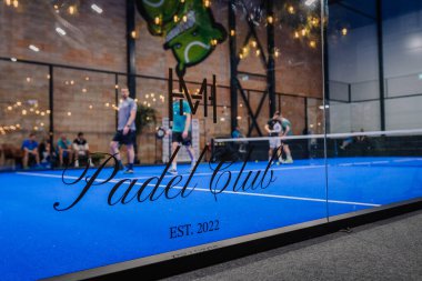 Riga, Letonya - 5 Nisan 2025 - Padel Club EST 'in yer aldığı cam duvardan görülebilen oyuncular ve izleyicilerle kapalı bir turnuva sahnesi. 2022 logosu ve modern ışıklandırma