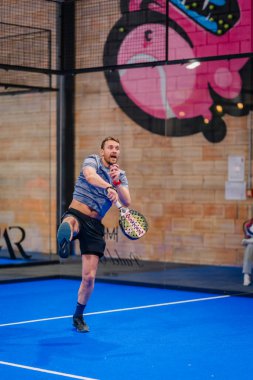 Riga, Letonya - 5 Nisan 2025 - Gri tişörtlü erkek padel oyuncusu, arka planda duvar yazısı olan mavi bir iç sahada düzenlenen bir padel turnuvasında güçlü bir hava saldırısını tamamladı..