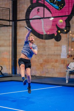 Riga, Letonya - 5 Nisan 2025 - Erkek padel oyuncusu, arka planda renkli grafiti ve tuğla duvar resmi olan mavi bir sahada bir padel turnuvası sırasında topu parçalamak için havaya sıçradı.