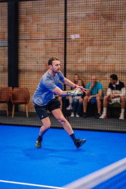 Riga, Letonya - 5 Nisan 2025 - Gri tişörtlü ve siyah şortlu odaklanmış erkek padel oyuncusu, kapalı alanda oynanan bir turnuva sırasında geri şuta hazırlanıyor..