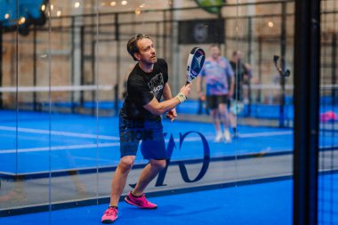Riga, Letonya - 5 Nisan 2025 - Siyah Adidas gömlekli ve pembe ayakkabılı erkek Padel turnuvası sırasında cam duvarların arkasındaki mavi kortta dönüş için hazırlanıyor.