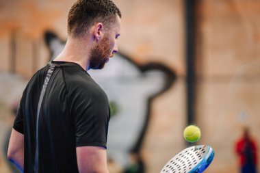 Riga, Letonya - 5 Nisan 2025 - Siyah tişörtlü bir erkek oyuncunun bir padel turnuvası sırasında raketinin etrafında uçuşan topu yakından izlemesi, arka plandaki saha ve duvarı bulanıklaştırması.