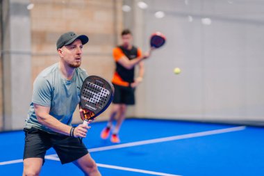 Riga, Letonya - 5 Nisan 2025 - Padel turnuvası sırasında iki erkek oyuncu dövüşüyor, biri hazır pozisyonda, diğeri ise kapalı mavi bir sahada atış yapıyor..