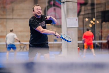 Riga, Letonya - 5 Nisan 2025 - Siyah kıyafetli odaklanmış erkek oyuncu, kapalı bir mavi sahada düzenlenen bir Padel turnuvası sırasında ters vuruş yaptı..