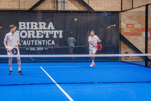 Riga, Letonya - 5 Nisan 2025 - Beyazlar içinde iki erkek padel oyuncu, arka planda markalaşma ile birlikte kapalı bir sahada mavi bir maçta mücadele ediyor..
