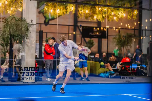 Riga, Letonya - 5 Nisan 2025 - Beyaz giyimli erkek padel oyuncusu, arka planda seyircilerin bulunduğu bir Padel turnuvası sırasında canlı bir iç sahada forehand atışı yaptı..