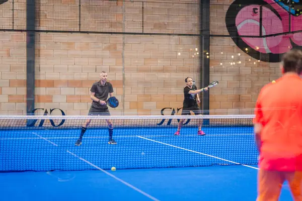 Riga, Letonya - 5 Nisan 2025 - İki erkek Padel turnuva maçında kalede hazırlanırken, turuncu giyen bir rakip karşı sahadan şut atıyor..