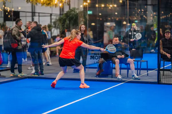 Riga, Letonya - 5 Nisan 2025 - Kırmızı tişörtlü bayan padel oyuncusu, arka planda seyirci ve oyuncuların yer aldığı bir Padel turnuvasında tekrar atış yapmak için hamlesini yapıyor..