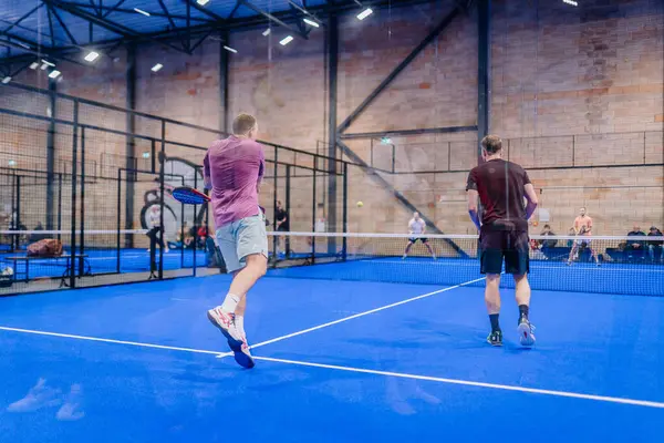 Riga, Letonya - 5 Nisan 2025 - Oyuncular dinamik bir iç saha padel turnuvasında yarışıyor, bir salıncakta atlarken diğerleri geri dönüş için hazırlanıyor, arka planda seyirciler izliyor.