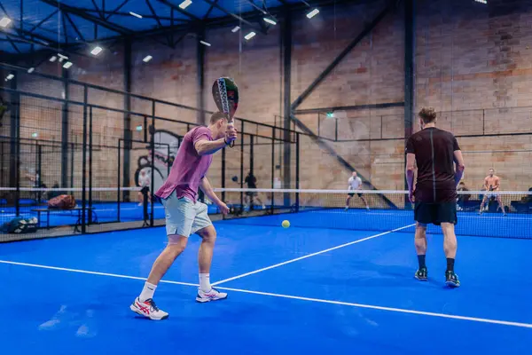 Riga, Letonya - 5 Nisan 2025 - Padel turnuvası maçında çarpışan iki adam, modern bir kapalı alan spor tesisi içinde canlı bir mavi korta ters vuruş hazırlıyorlar..