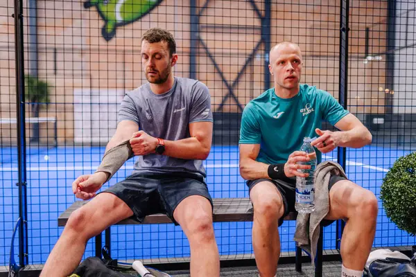 Riga, Letonya - 5 Nisan 2025 - Padel turnuvası sırasında iki erkek padel oyuncusu bir bankta dinleniyor, biri kollarını düzeltiyor, diğeri ise arka planda mavi bir iç saha ile içme suyu içiyor..