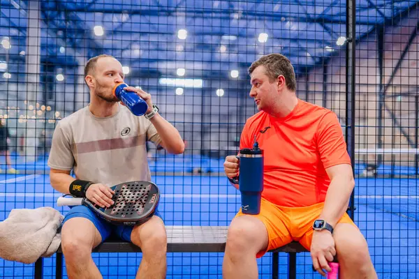 Riga, Letonya - 5 Nisan 2025 - İki Padel oyuncusu kapalı bir sahada düzenlenen bir turnuva sırasında su şişeleriyle su içerek yedek kulübesinde mola verdi.