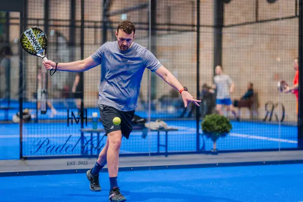 Riga, Letonya - 5 Nisan 2025 - Gri tişörtlü erkek padel oyuncusu, kapalı alanda düzenlenen ve arka planda oyuncuların yer aldığı bir turnuva sırasında düşük topa vurmaya hazırlanıyor..