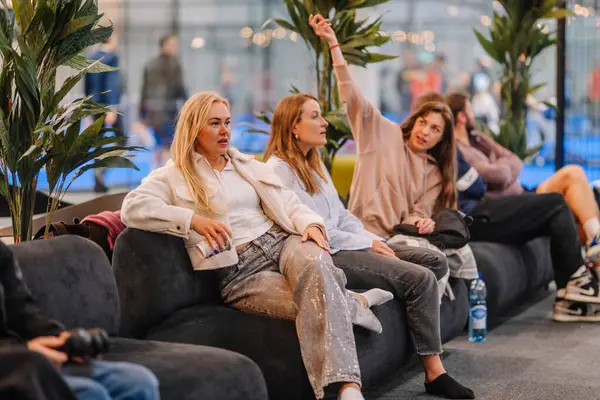 Riga, Letonya - 5 Nisan 2025 Modern bir padel kulübünün lobisinde bir kanepede oturan kadınlar cam duvardan padel turnuvasını izleyip sohbet ediyorlar..