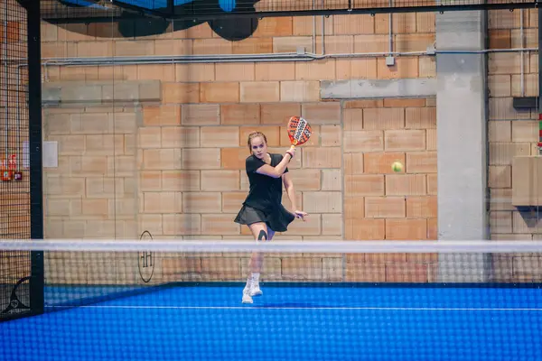 Riga, Letonya - 5 Nisan 2025 - Siyah kıyafetli kadın padel oyuncusu kapalı mavi bir sahada bir ön atış sırasında arka planda ağ ve tuğla duvarla vuruyor.