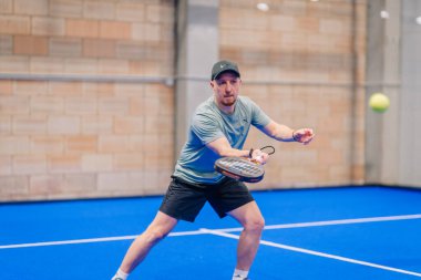 Riga, Letonya - 5 Nisan 2025 - Gri tişörtlü ve siyah şortlu erkek padel oyuncusu, kapalı mavi kortta düzenlenen bir turnuva sırasında şuta karşılık vermeye hazırlanıyor..