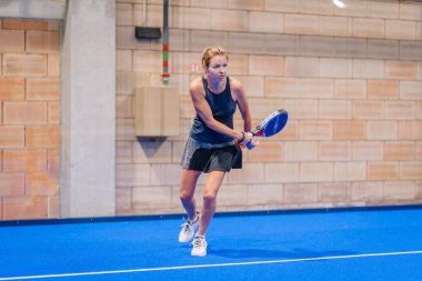 Riga, Letonya - 5 Nisan 2025 - Odaklanmış siyah kıyafetli kadın padel oyuncusu, kapalı mavi bir kortta, tuğla duvarlı bir padel turnuvası sırasında iki elle ters vuruş yapmaya hazırlanıyor..