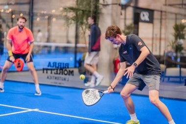 Riga, Letonya - 5 Nisan 2025 - Padel turnuvası sırasında iki erkek oyuncu, biri mavi iç sahada siyah ve kırmızı spor giyim giyerek geri dönüş atışına hazırlanıyor..