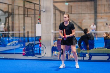 Riga, Letonya - 5 Nisan 2025 - Odaklanmış siyah kıyafetli bayan oyuncu, kapalı alandaki mavi sahada seyirciler cam duvarın arkasında otururken Padel turnuvasında servis almaya hazırlanıyor..