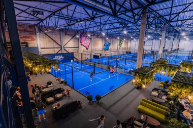 Riga, Letonya - 5 Nisan 2025 - Birden fazla sahası olan kapalı padel turnuva salonu, oyuncular hareket halinde ve yeşil, ışıklar ve seyircili canlı bir merkez dinlenme alanı.