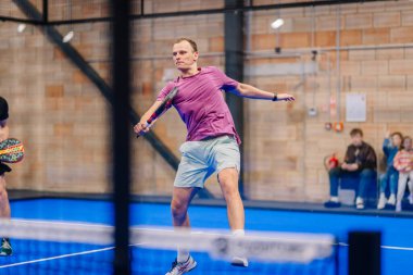 Riga, Letonya - 5 Nisan 2025 - Mor gömlekli ve açık renk şortlu bir adam Padel turnuvası sırasında mavi iç sahada raket sallıyor..