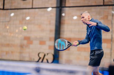 Riga, Letonya - 5 Nisan 2025 - Mavi tişörtlü ve siyah şortlu adam kapalı sahadaki bir padel maçında topa vurmaya odaklandı, elinde siyah raket, arkasında tuğla duvar vardı.