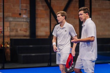 Riga, Letonya - 5 Nisan 2025 - Beyaz spor giyimli iki erkek oyuncu kapalı alanda oynanan bir turnuva maçında konuşuyor, ellerinde kırmızı padel raketi, tuğla duvar arkası.
