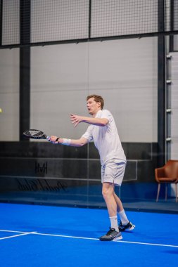Riga, Letonya - 5 Nisan 2025 - Kapalı bir sahada padel oynayan adam, siyah raketle bir ralli sırasında, mavi bir yüzey üzerinde beyaz spor giysisi ve siyah Nike ayakkabıları giymişti..