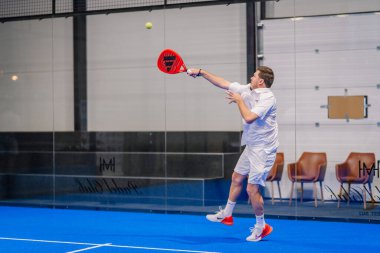 Riga, Letonya - 5 Nisan 2025 - Padel oynayan, kırmızı raketle orta sahada top oynayan, beyaz giyimli, turuncu ayakkabılı, arka planda modern spor yapan adam.